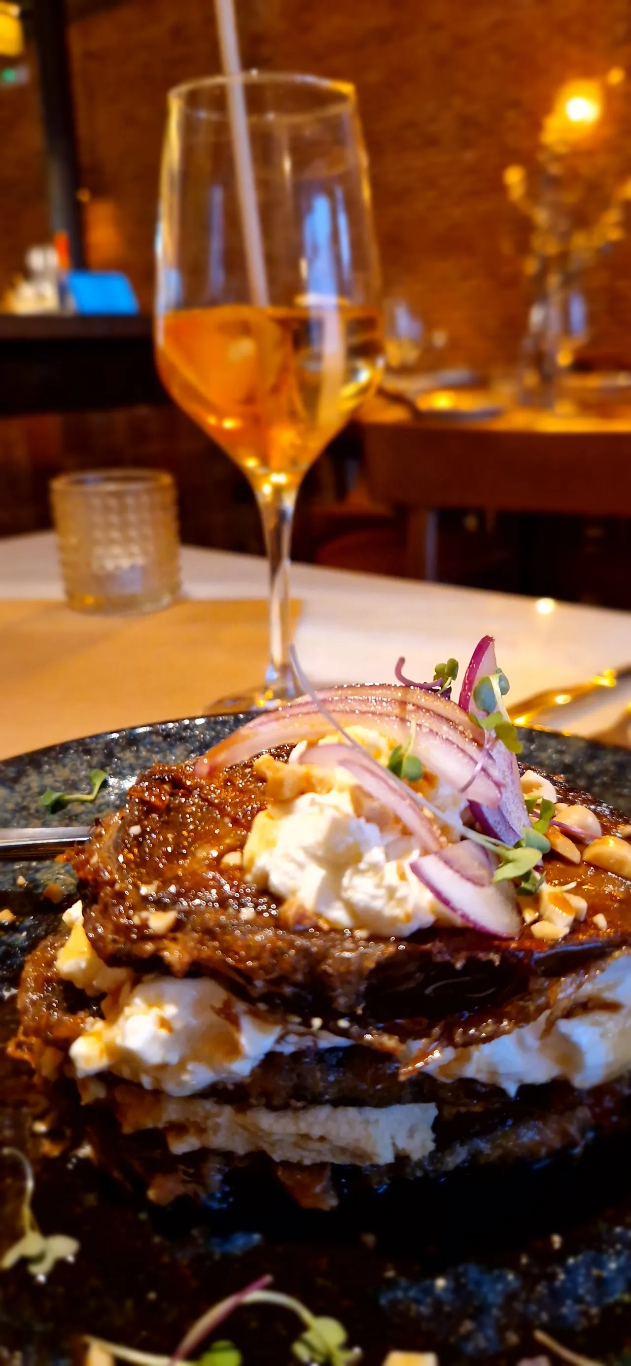 A stacked tower dish with layers of toast, egg, and toppings beside a glass of wine