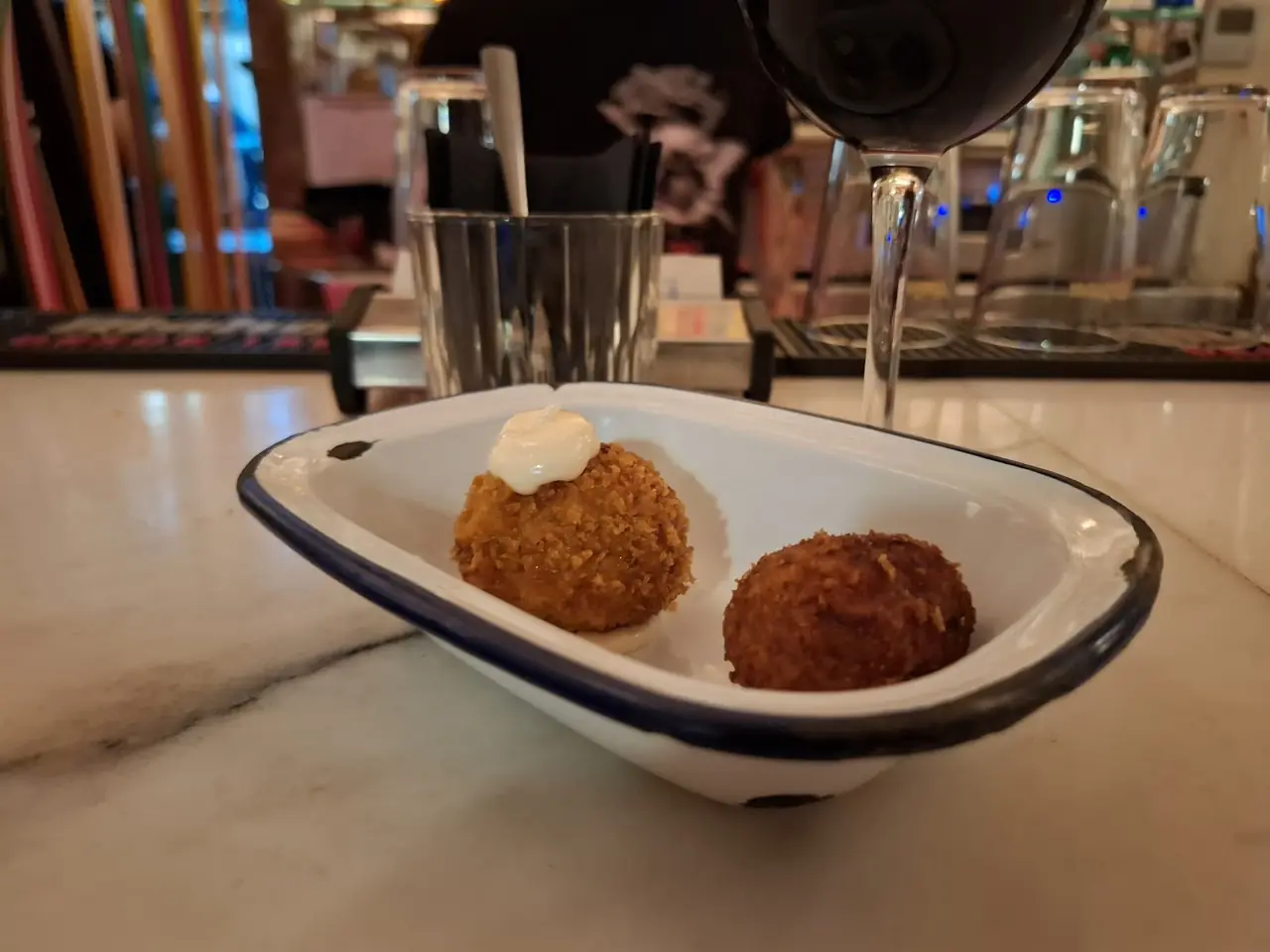 Two breaded fried croquettes in a small dish, one topped with sauce, glass of red wine beside