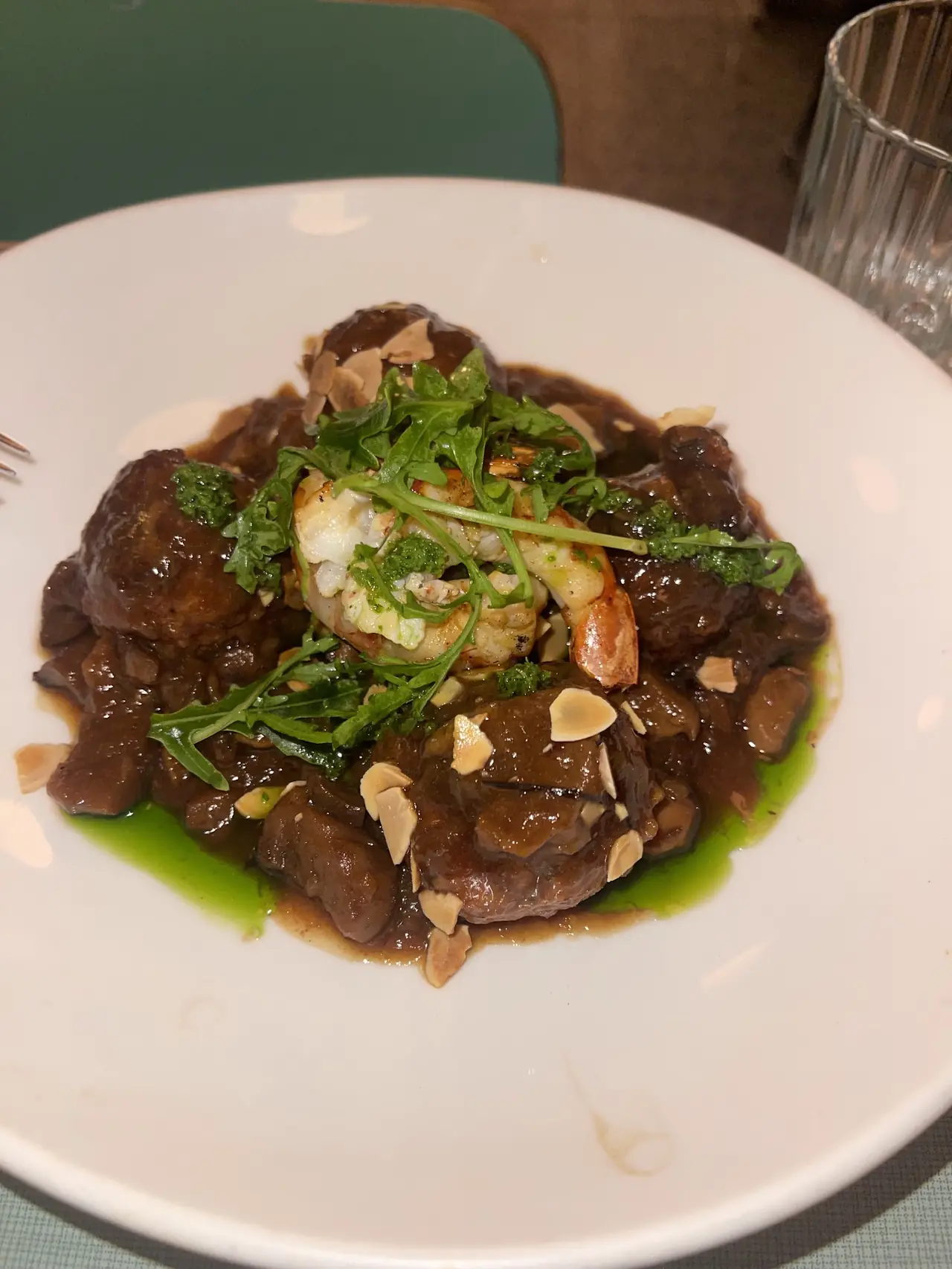 Braised meat stew with a grilled prawn, arugula, sliced almonds and green oil in a white bowl