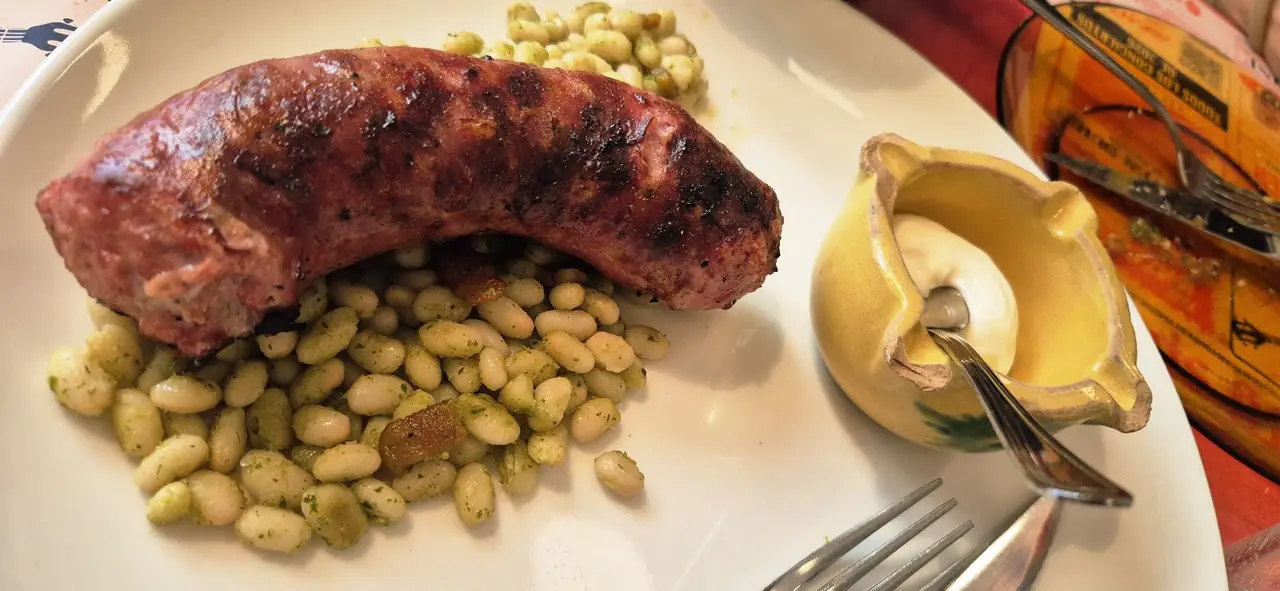 Grilled sausage over white beans in green sauce on a plate, small pot of mayonnaise beside