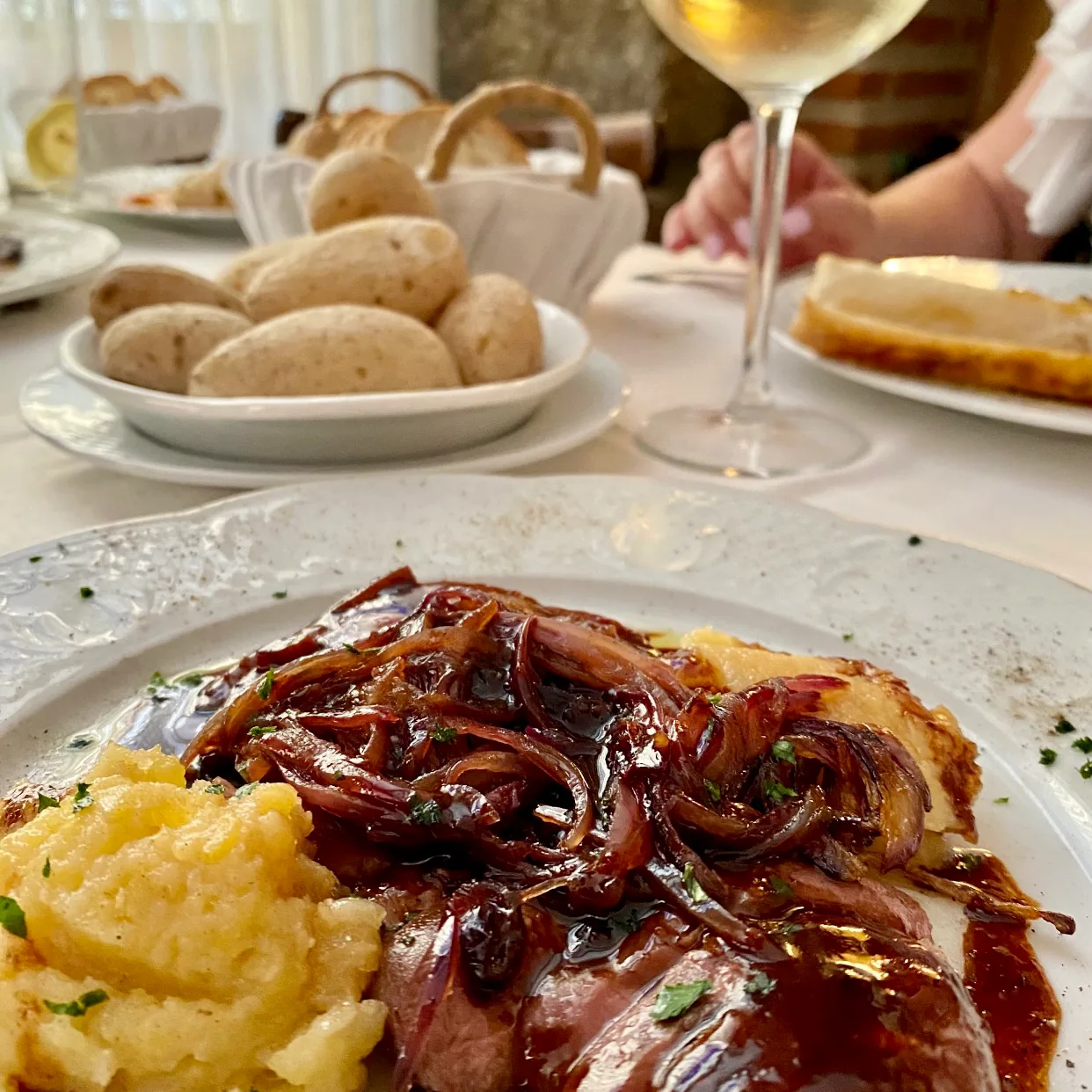 A plate of roasted meat with mashed potato and glazed onions, served with bread rolls and wine