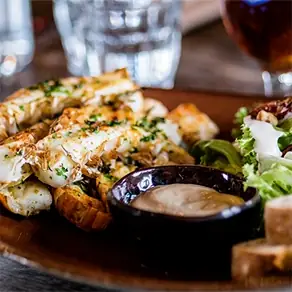 Wooden board with golden fried strips of fish or chicken, a bowl of pale dipping sauce, green salad and bread