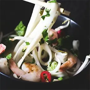 A dark bowl of noodle soup with prawns, sliced red chilli, herbs and bean sprouts, chopsticks lifting white noodles from the bowl