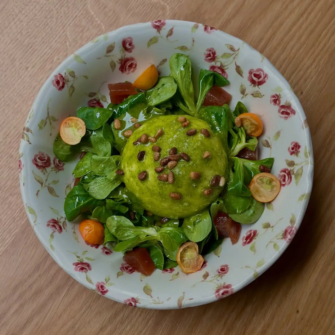 Overhead shot of a round green dish topped with pine nuts, served with mache salad, tomato, and kumquat slices on a floral-patterned plate