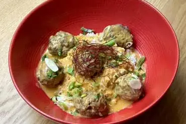 Bowl of meatballs in a creamy sauce garnished with fried leek strands and spring onion, in a red bowl