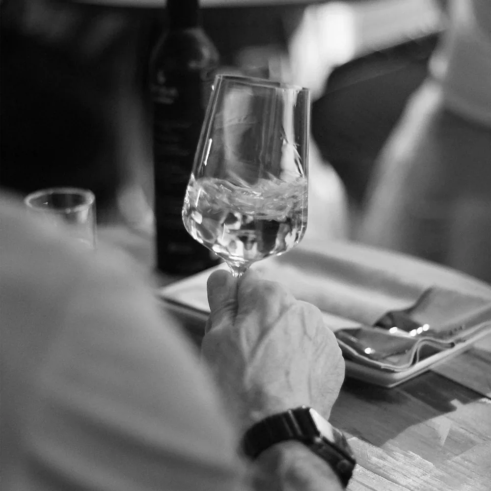 A hand swirling wine in a tasting glass at a laid table in black and white, an open bottle standing behind and cutlery on a napkin alongside
