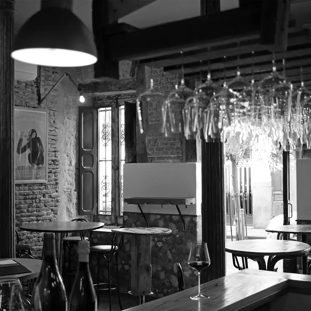 La Fisna's room in black and white, exposed brick and stone, wooden beams, glasses hung upside down from an overhead rack, small cafe tables and bentwood chairs, and a poured glass of red wine on the bar in the foreground