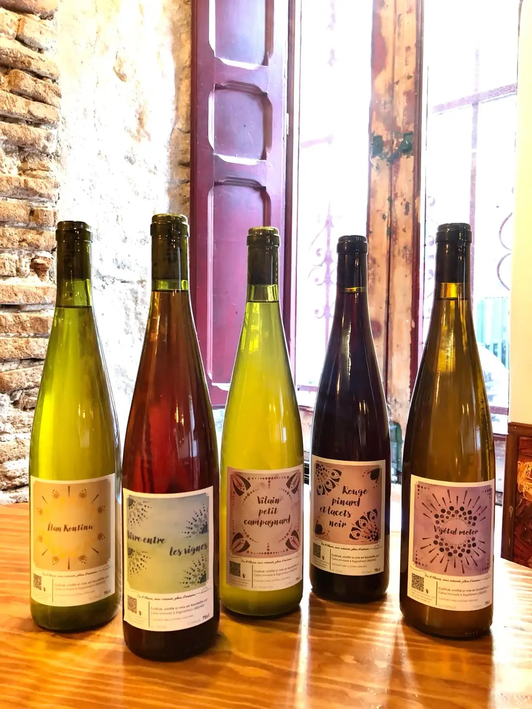 Five natural wine bottles with hand-lettered labels lined up by a window