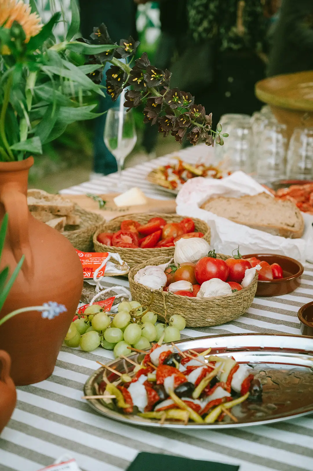Sliced heirloom tomatoes, garlic bulbs, green grapes, crusty bread, and cheese laid out on a striped tablecloth at La Font del Gat
