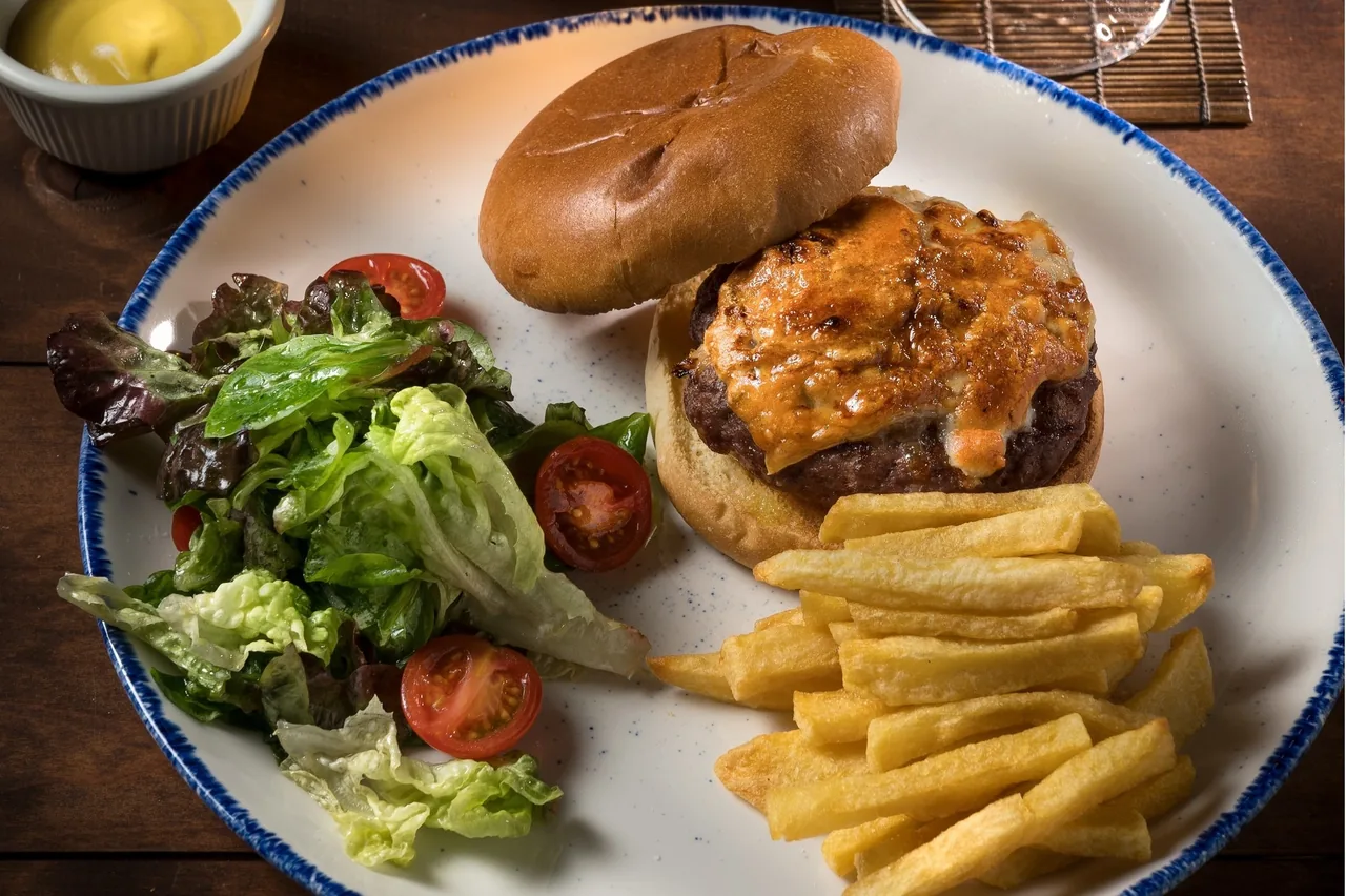 A burger with a cheese-topped patty on a white plate with fries, salad and a small pot of mustard