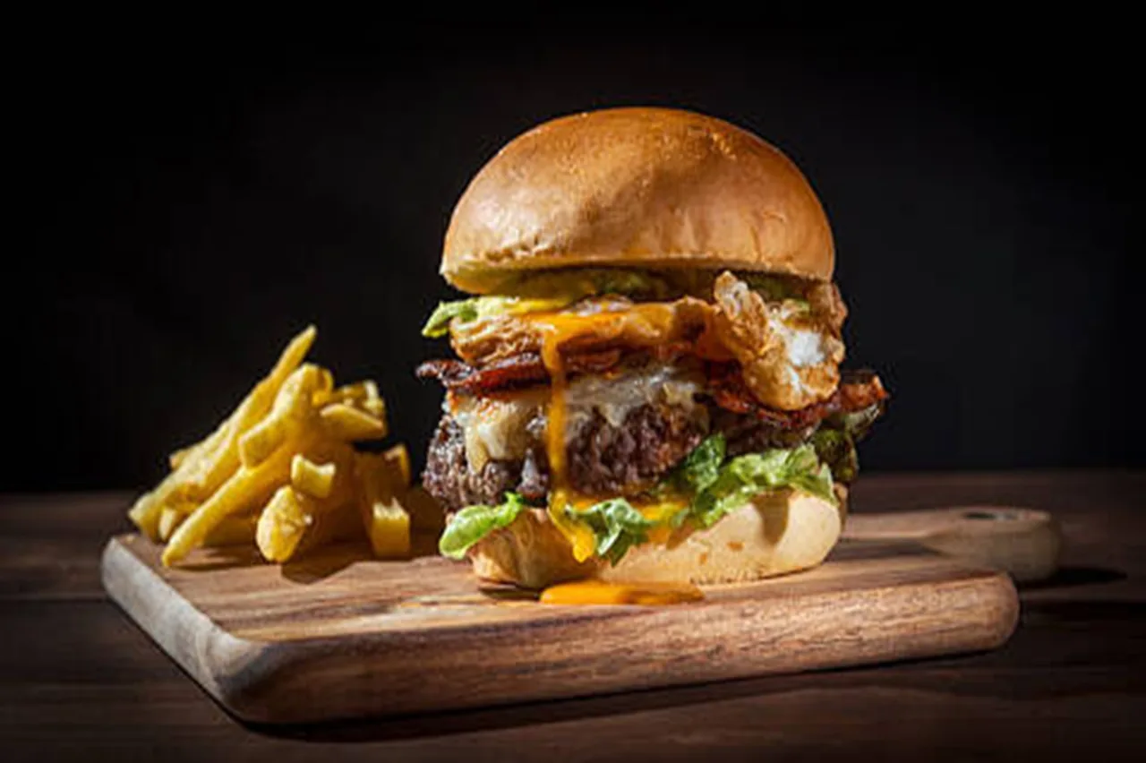 A tall burger with a fried egg, bacon and lettuce on a wooden board beside fries