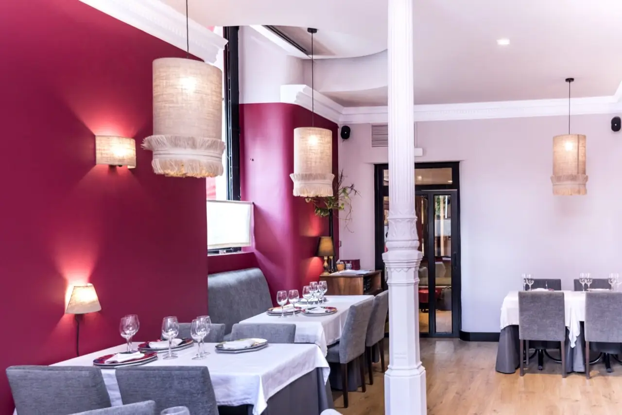 Dining room with burgundy accent walls, white pillar, linen pendant lamps and tables set with white cloths, wine glasses and colourful chargers