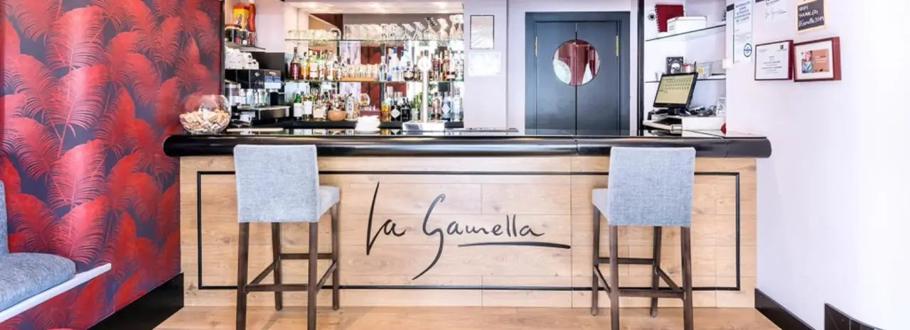 Bar counter with the name La Gamella scripted on its wood-panelled front, two grey bar stools, back bar shelves of spirits and glassware, red palm-print wallpaper to the left