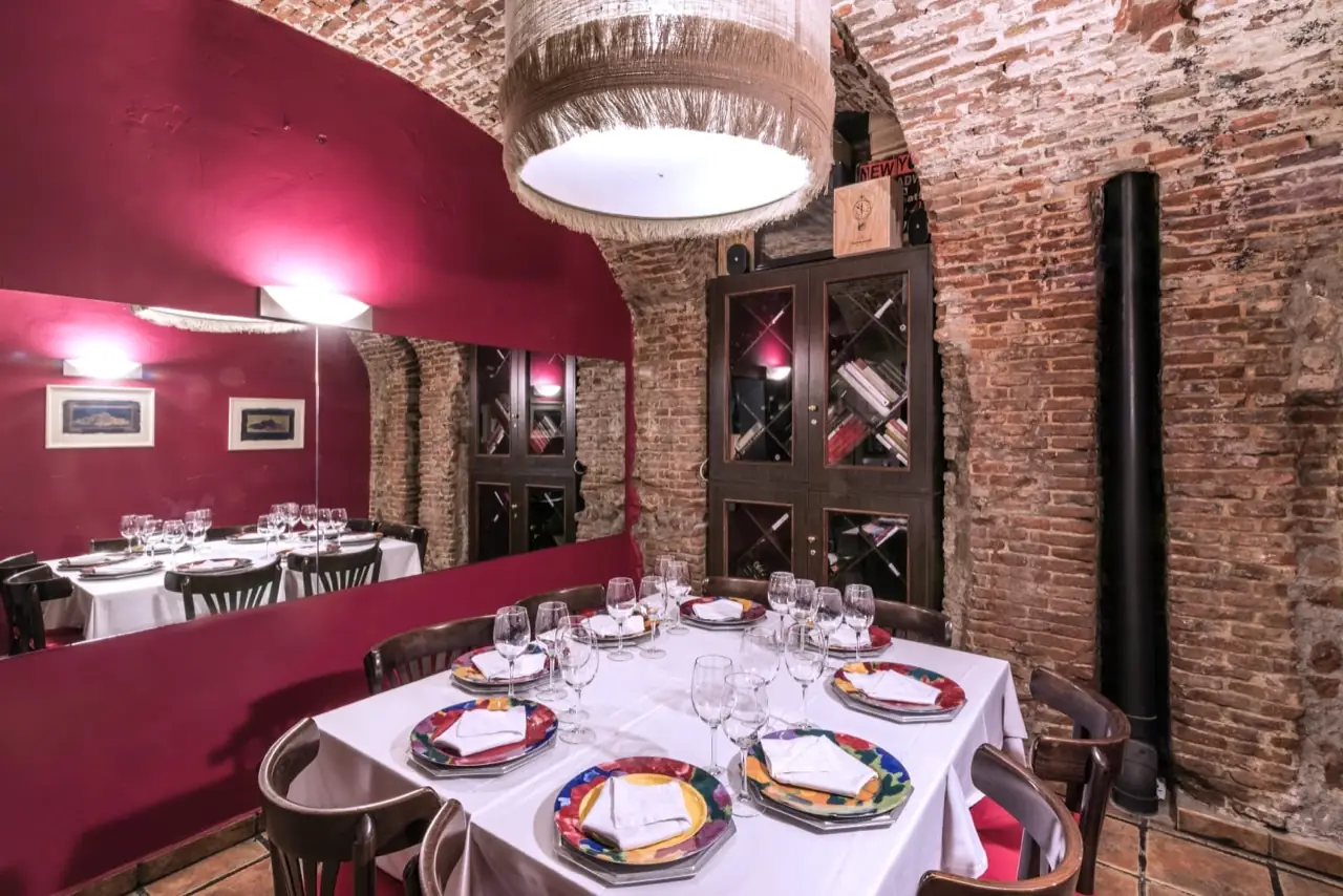 Brick-vaulted dining room with a burgundy wall, large mirror, glass-fronted wine cabinet and a table set with white cloth and multicoloured plates