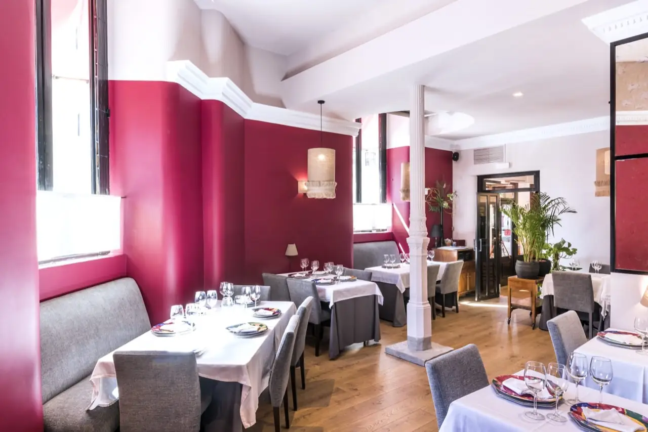 Bright dining room with burgundy and white walls, a slim white pillar, wooden floor and several tables set with white linens and colourful chargers