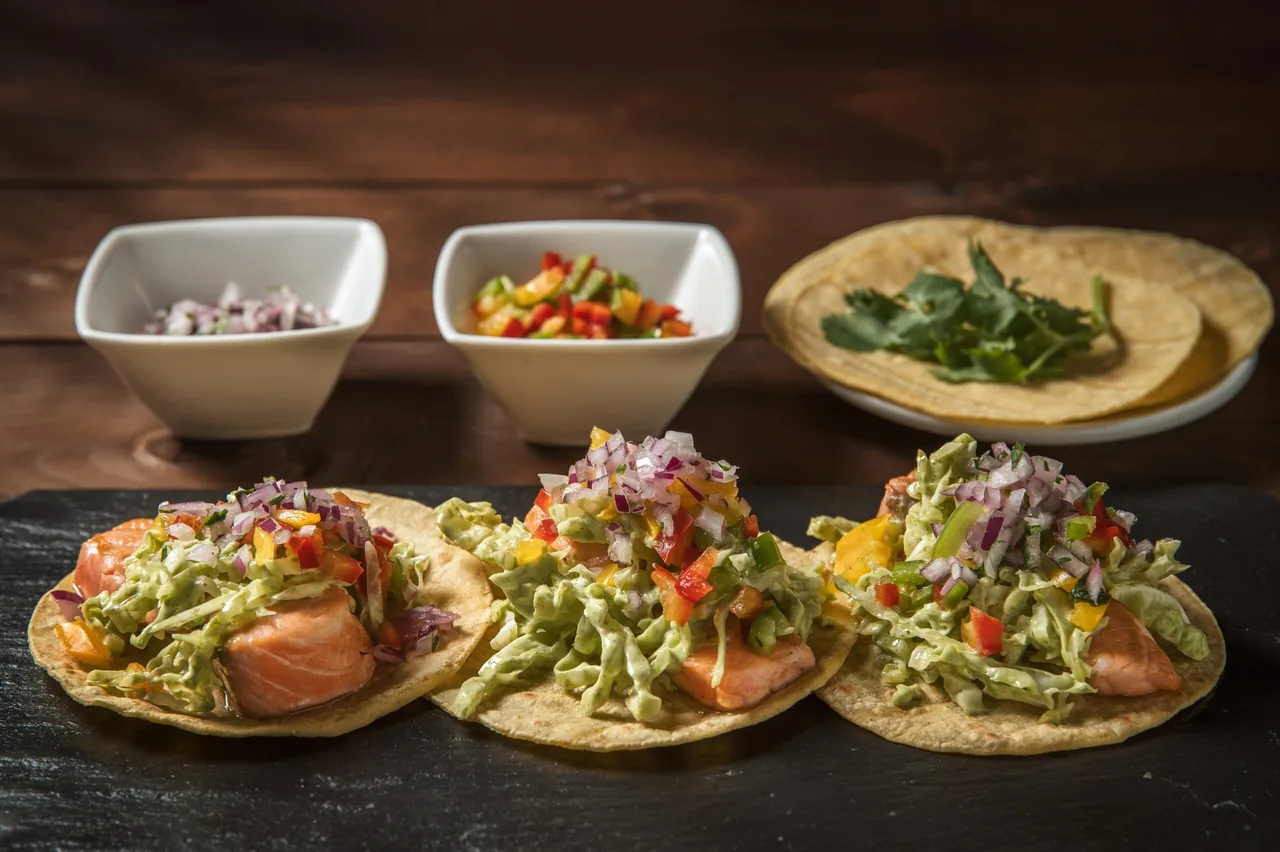 Three tacos topped with shredded green salad, chopped onion and pepper on a slate board, with small bowls and a stack of tortillas behind