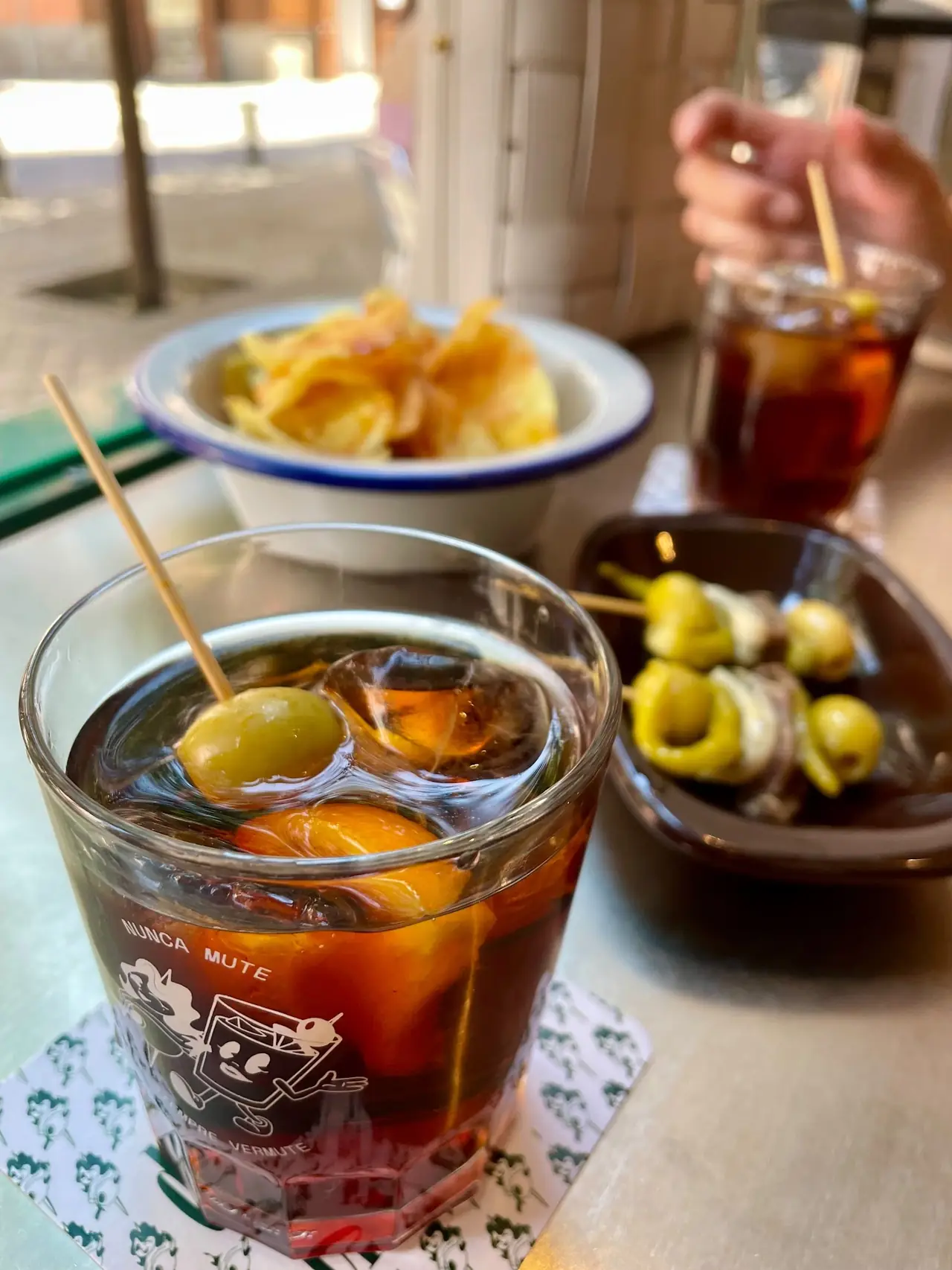 Glass of vermut on ice with an olive garnish beside chips and gilda skewers