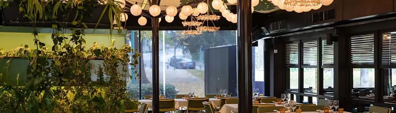 Dining room with paper lantern string lights and beaded chandeliers, dark wood columns, tables visible near large windows onto the street, and trailing plants on the left.