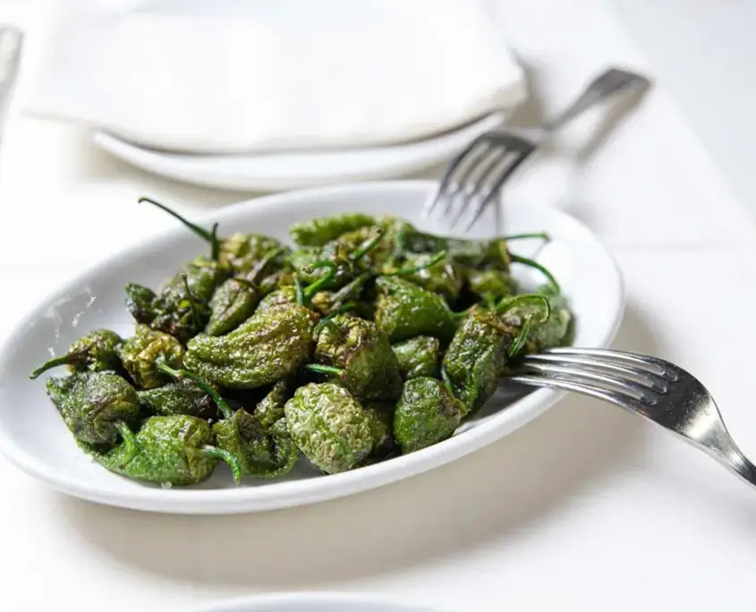 White oval plate of blistered padron peppers with two forks resting across it on a white tablecloth.
