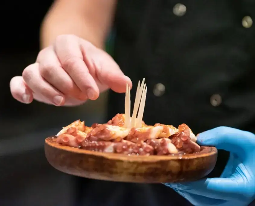 Gloved hands plating chopped octopus in paprika sauce on a wooden dish, adding toothpicks.