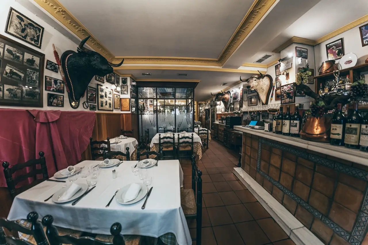 Narrow traditional dining room with white-clothed tables, mounted bull heads, framed bullfighting photographs, and a bar lined with wine bottles and copper pots