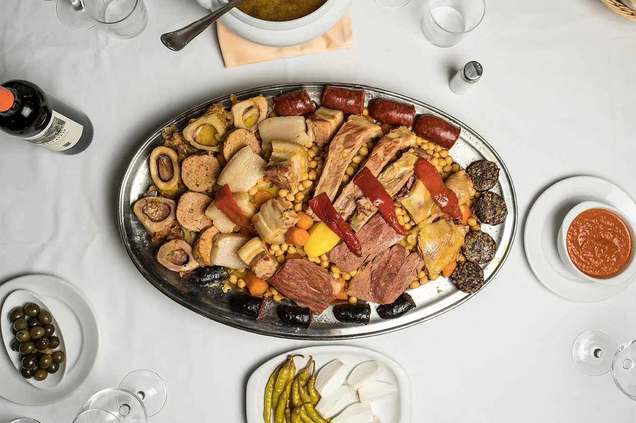 An oval metal platter of sliced boiled meats, chorizo, morcilla and red peppers seen from above on a white tablecloth