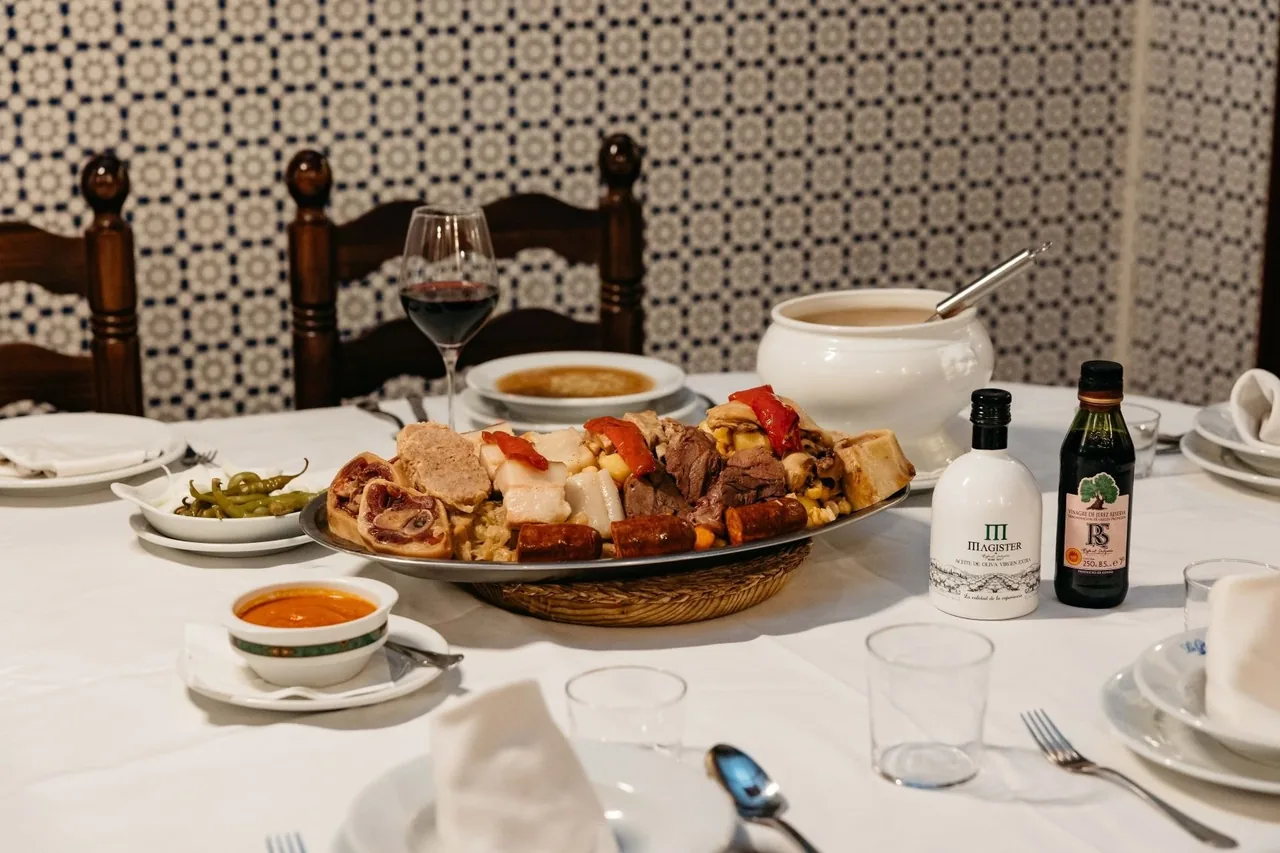A platter of boiled meats and chorizo on a white-clothed table with a soup tureen and a glass of red wine in front of a tiled wall