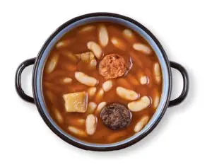 Fabada asturiana: a black two-handled pot of creamy white beans in reddish broth with slices of chorizo and morcilla, shot overhead on white