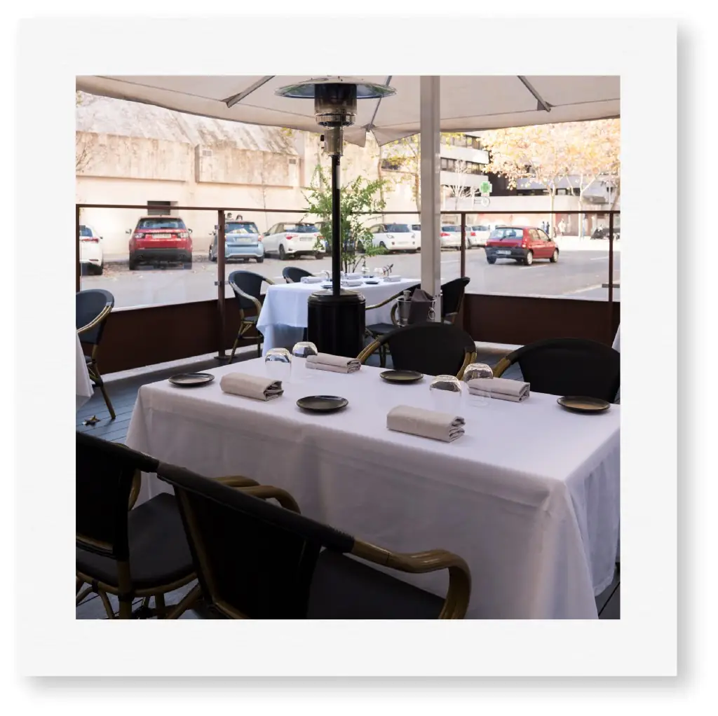 The venue's covered street terrace: white-clothed tables set with dark plates, folded napkins and glasses under a canopy with a patio heater, parked cars beyond the railing