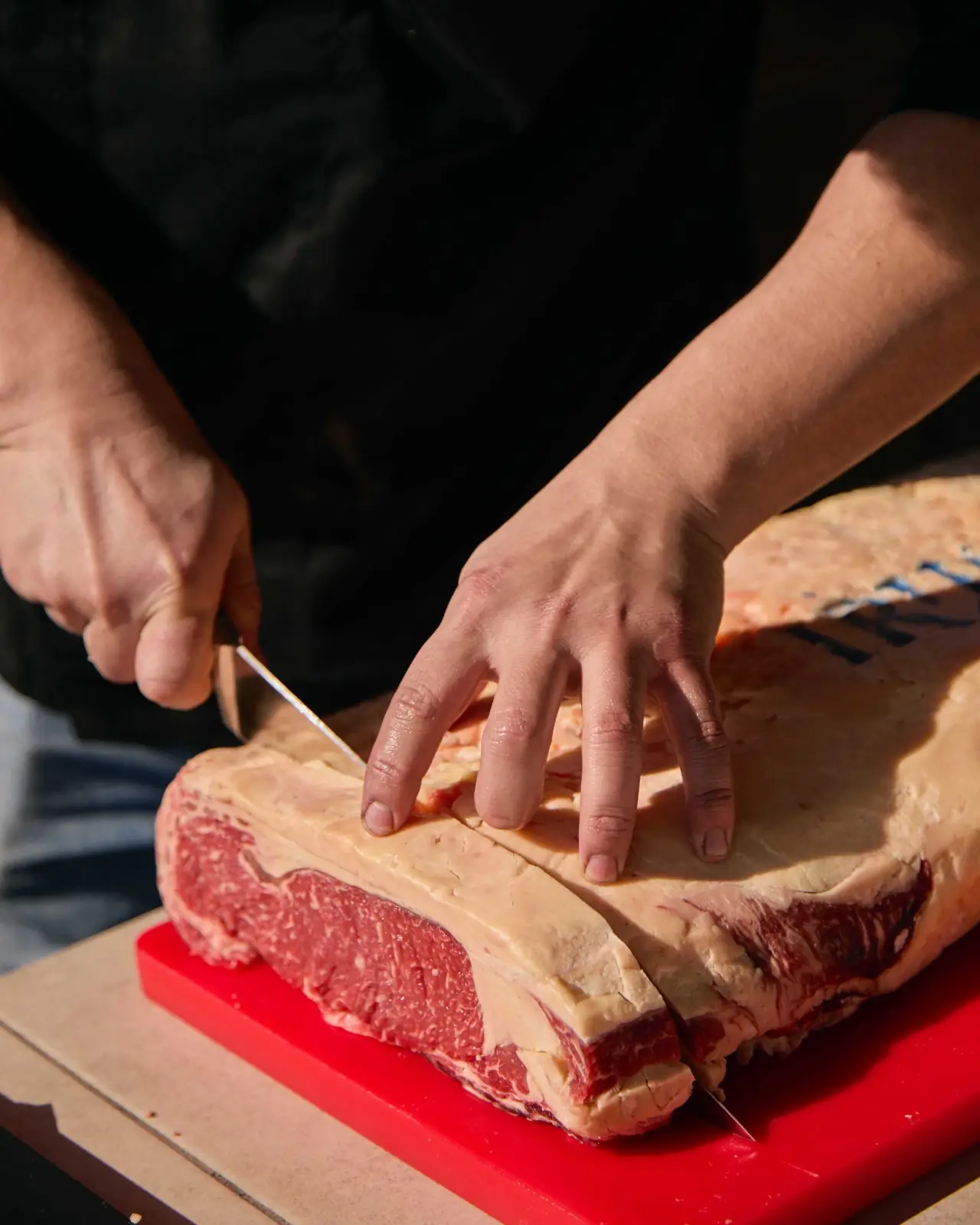 Hands slicing a large marbled dry-aged beef loin with a knife on a red cutting board