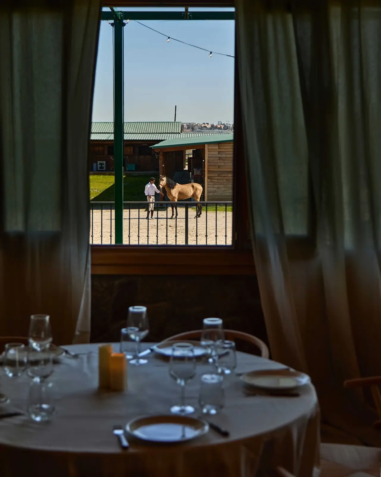 A set round dining table in the foreground with a window view onto stables and a handler leading a horse