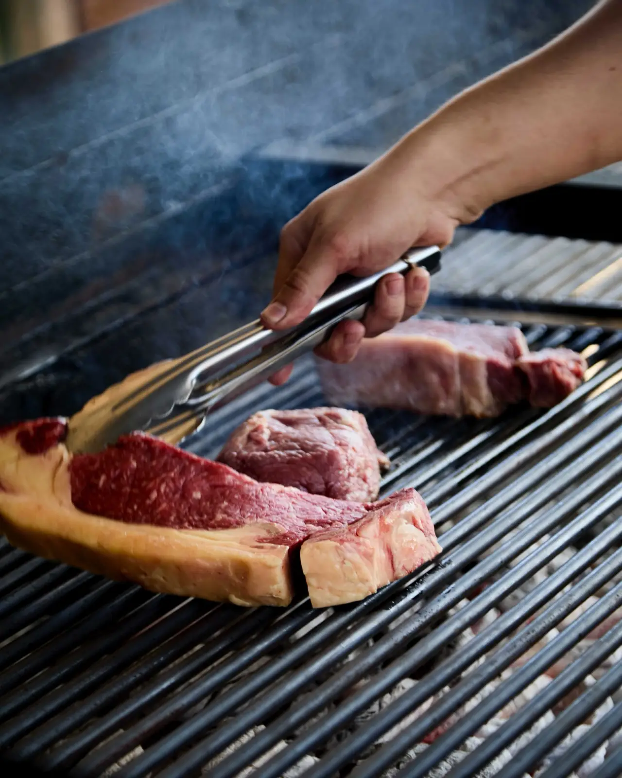 A hand using tongs to grill large cuts of steak over a smoking grill