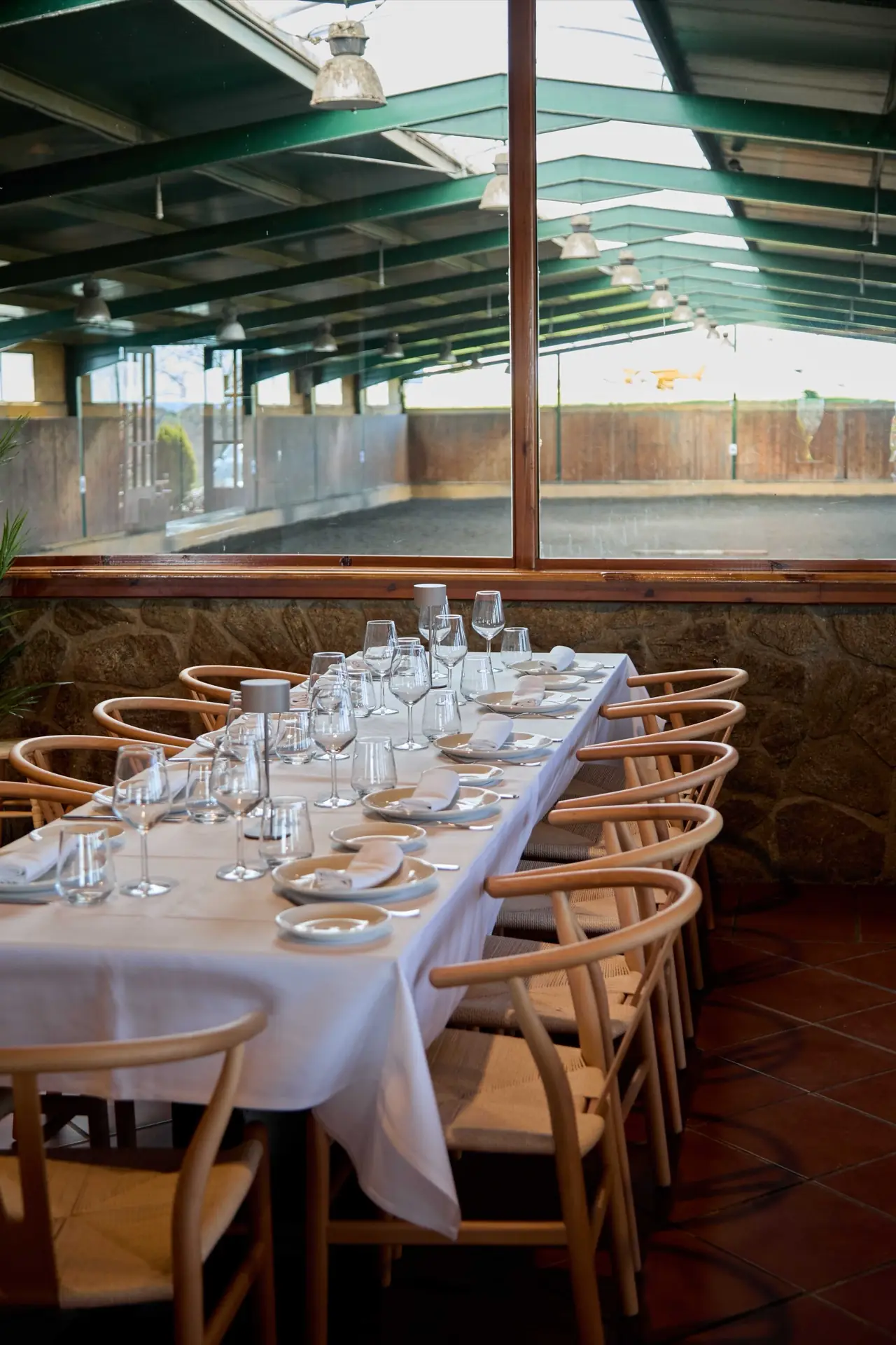 Long white-linen table set with wine glasses and wishbone chairs beside a glass wall overlooking the riding arena