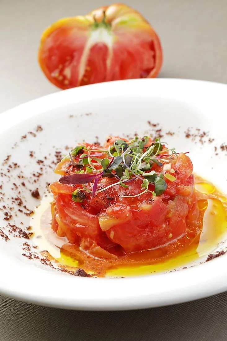 Chopped red tomato topped with micro herbs and dark crumbs in a white bowl
