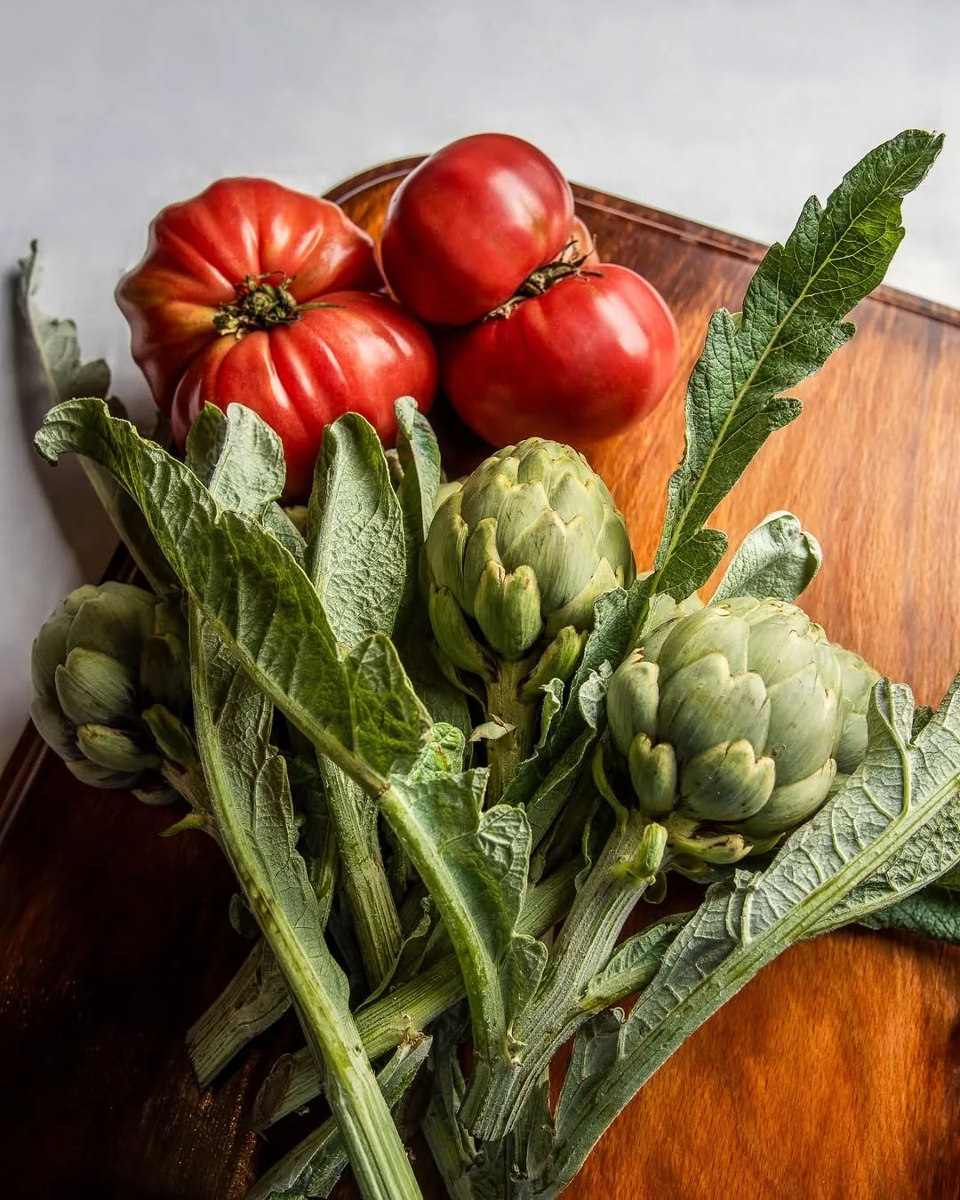 Fresh artichokes, leafy stems and ripe red tomatoes on a wooden board