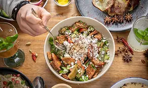 Overhead bowl of stew or grain dish topped with crumbled cheese and herbs, a hand holding a fork over it on a wooden table with side dishes and star anise