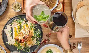Overhead table of Middle Eastern plates with two hands raising a mint drink and a glass of red wine over garnished dishes