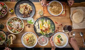 Overhead spread of many hummus bowls, salads, pita bread and mezze plates on a wooden table with several diners' hands reaching in