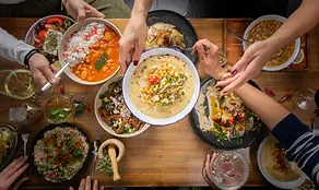 Overhead spread of hummus and rice dishes on a wooden table, hands sharing food, wine glasses and a mortar around the plates