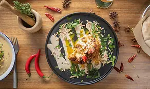 Black plate of topped hummus drizzled with olive oil and garnished with herbs, red chilies and a mortar on a wooden table