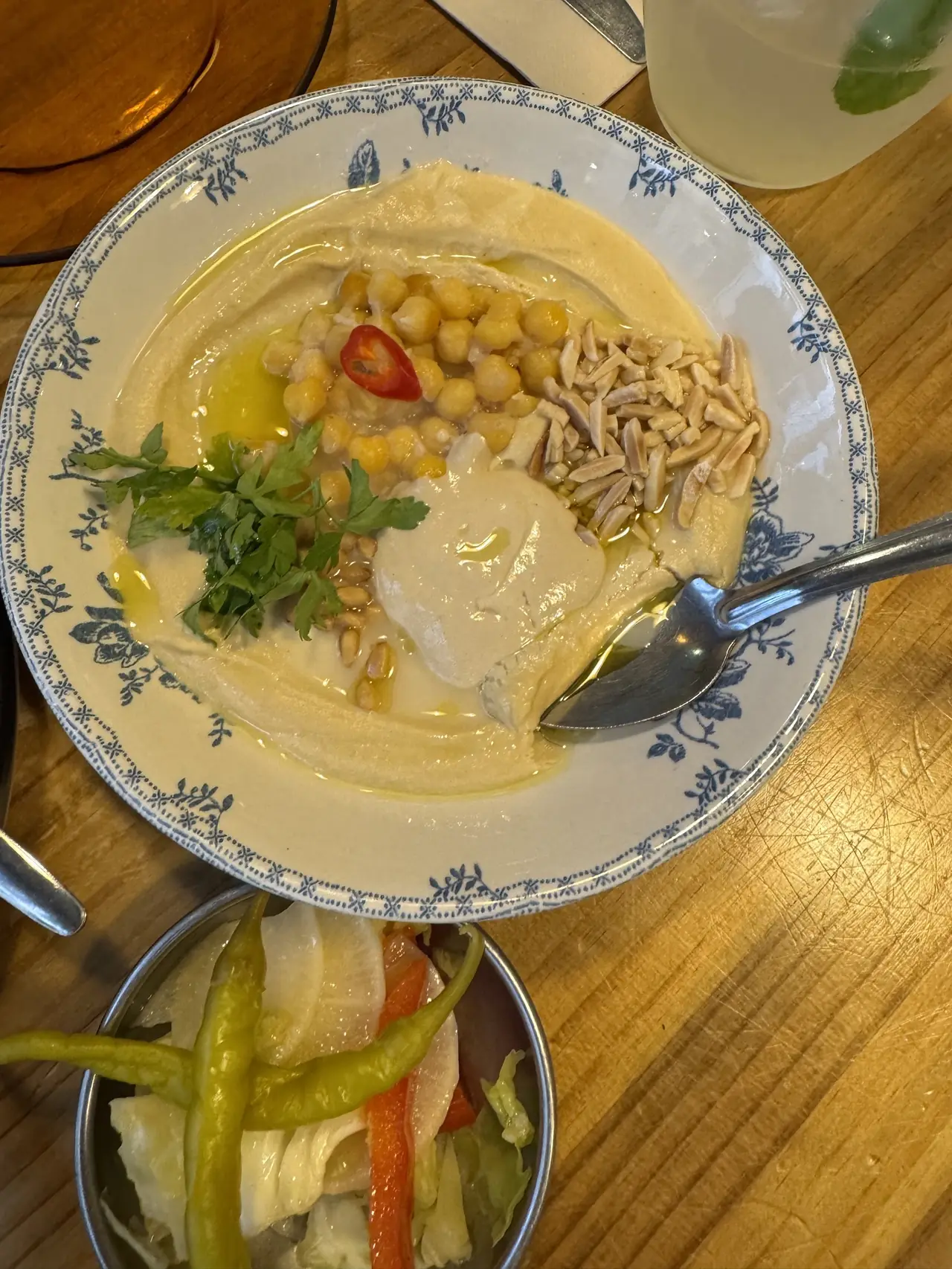 A bowl of hummus topped with chickpeas, almonds, herbs and a chilli, with pickles alongside