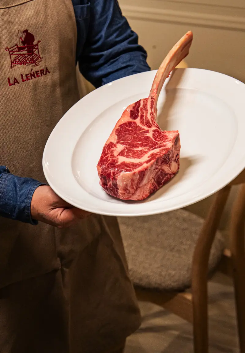 A person in a denim apron embroidered with the restaurant name holds a white plate with a large bone-in tomahawk steak.