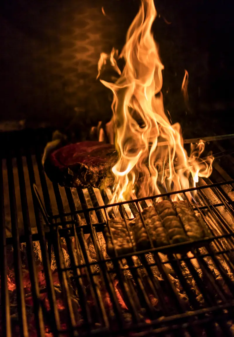 Large flames rising from a wood-fired grill with cuts of meat cooking on the grate below.