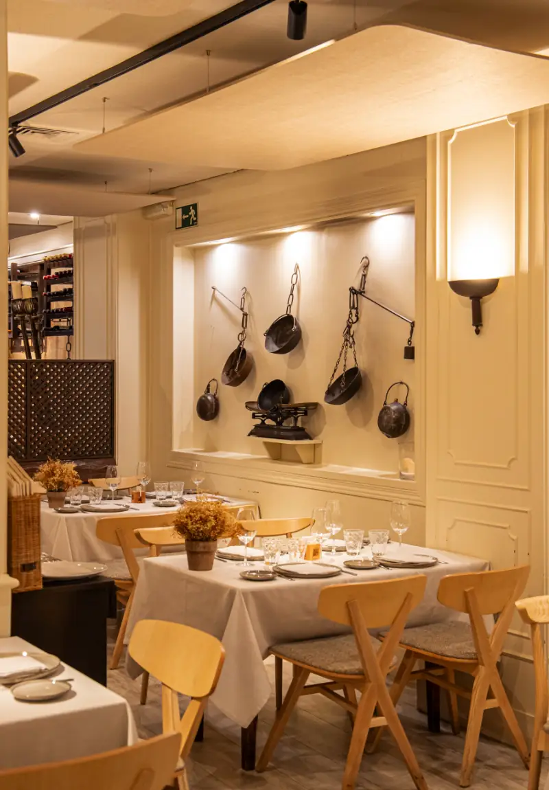 Dining room with wooden crossback chairs and set tables beneath a wall niche displaying antique hanging scale and cookware.