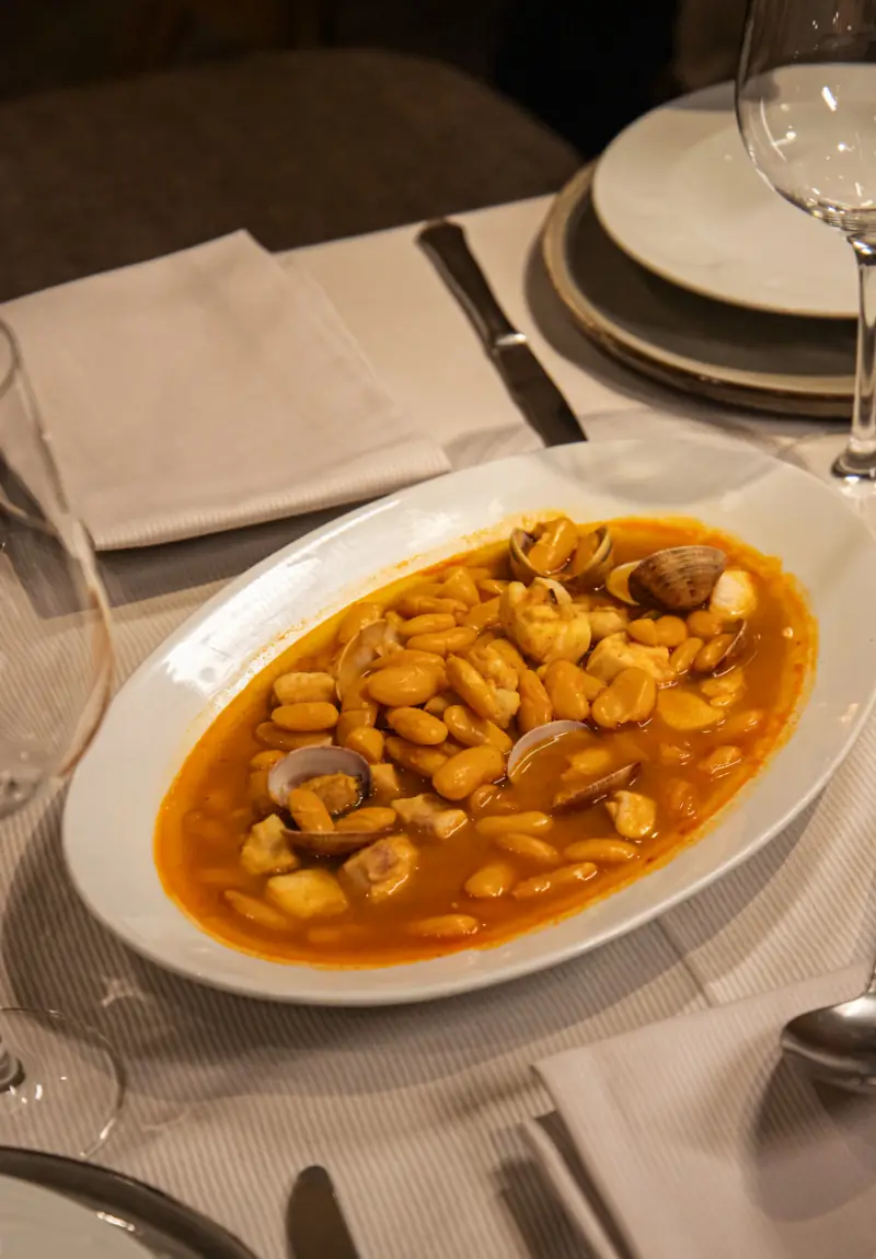 Oval white plate of white bean stew with clams and chunks of fish, set on a striped tablecloth beside a wine glass.