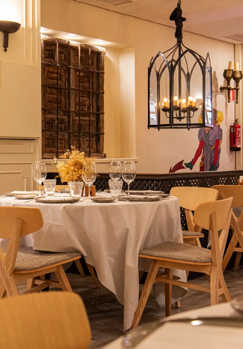 Dining room with a round white-clothed table set for a meal, wooden chairs, a gothic-style hanging light fixture and an antique wooden door panel on the wall.