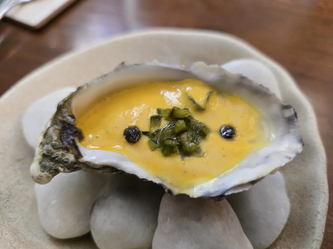 An oyster in its shell topped with a yellow sauce and capers on smooth stones