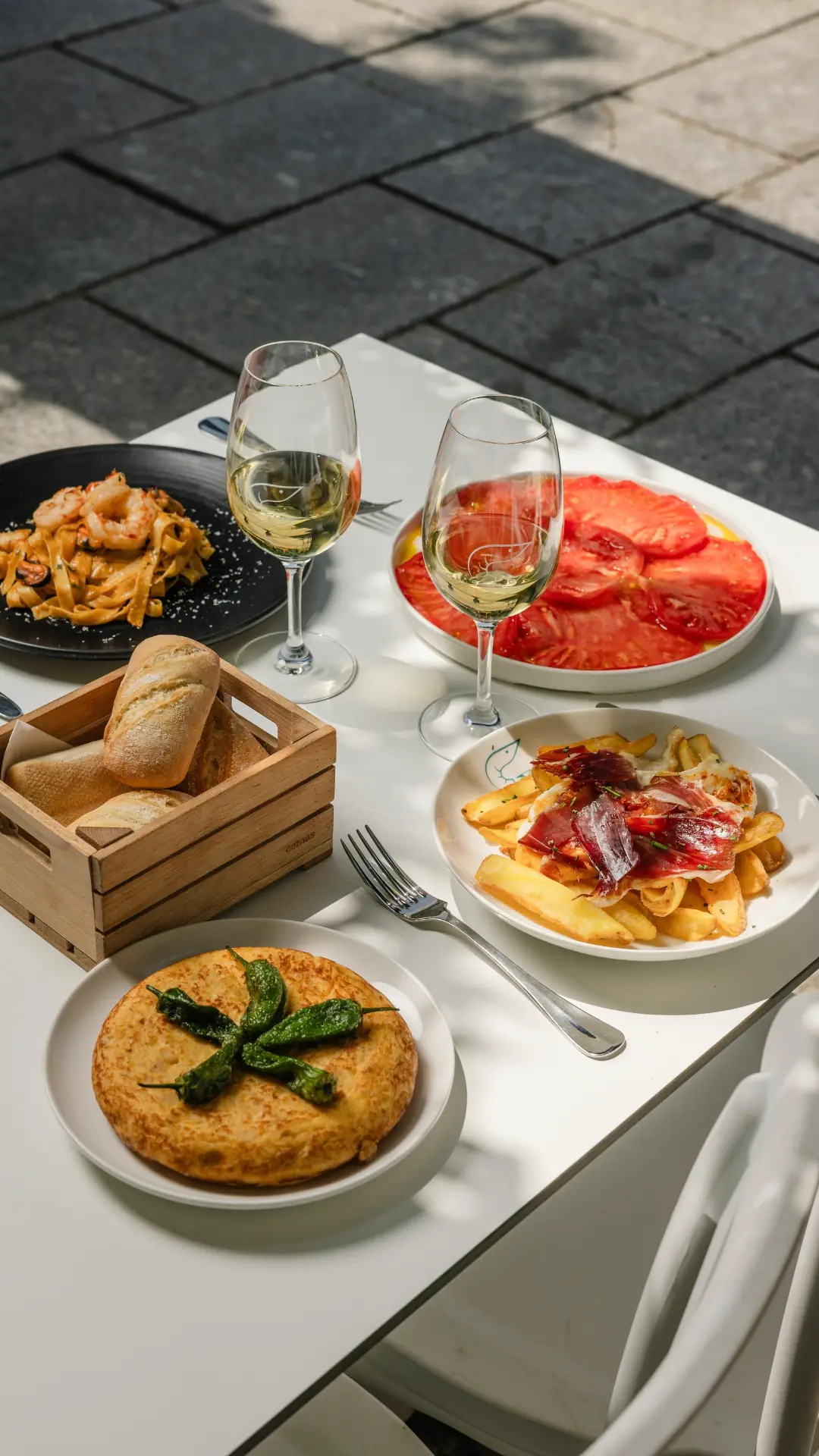 Outdoor table set with pasta, sliced tomatoes, patatas bravas with jamón, tortilla, bread, and two glasses of white wine