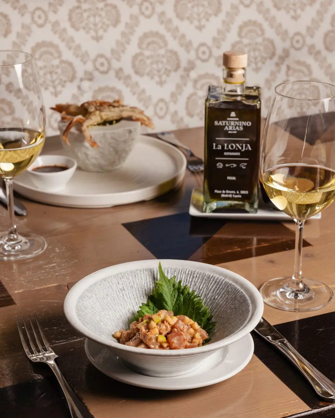 A tartare dish with shiso leaf on the table beside wine glasses, a fried seafood dish, and an olive oil bottle