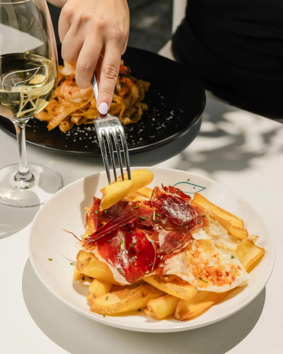 A hand with a fork over a plate of patatas bravas topped with jamón and fried egg, with pasta and wine in the background